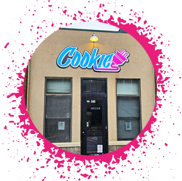 Tan storefront with brick trim that has a white, black, light blue, purple, and pink gradient sign that says 'Cookie' with an electrical plug attached to it.