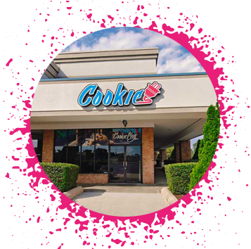 A tan and brick storefront with blue puffy clouds that has a sign over their door that says 'Cookie' with an electrical plug attached to it with large pictures of cookies in the window.