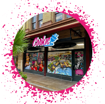 Tan building with neon pink, white, and blue sign that says 'Cookie' with an electrical plug attached to it. Brick building has large images of cookies and graffiti in the windows.