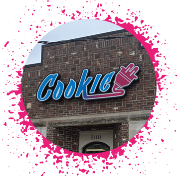 Blue, pink, and purple gradient sign says 'Cookie Plug' over a brick storefront with large windows with a hot pink spray paint border