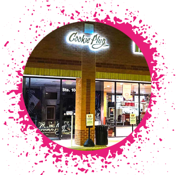 Brick storefront with a lit up sign that says 'Cookie Plug' that has a pink paint splash border around the outside.