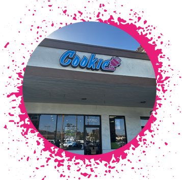 Beige storefront with a large blue, purple, and pink gradient sign that says 'Cookie' with an electrical plug attached to the end in graffiti letters over a window sign that says 'Now Hiring'.