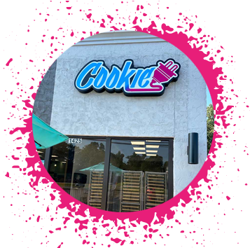 gray stone storefront with a blue, purple, and pink sign in graffiti type that says 'Cookie' with an electrical plug attached to it with cookie baking racks in the window.
