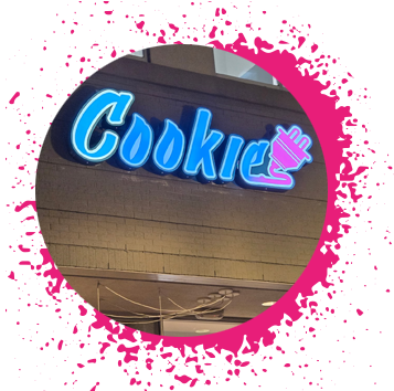 Blue, pink, and purple gradient sign says 'Cookie Plug' over a tan storefront with a pink paint splatter circular border.