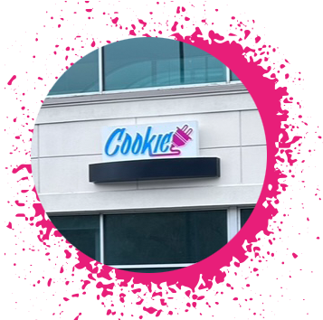 Blue, pink, and purple gradient sign says 'Cookie Plug' over a white brick storefront with large windows with a hot pink spray paint border