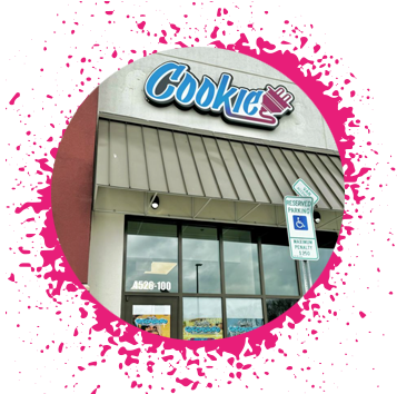 White and tan building with a large neon blue sign that says 'Cookie' with a hot pink electrical plug attached to it.