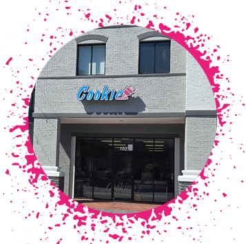 Gray brick storefront that has a blue and pink sign that says 'Cookie' with an electrical plug attached to the end of it.